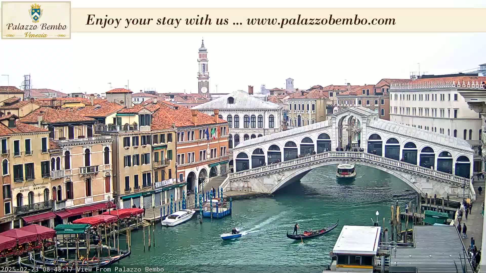A view of a canal in Venice on an overcast day, showing a bridge with people walking across it and gondolas in the water, with buildings lining the canal.