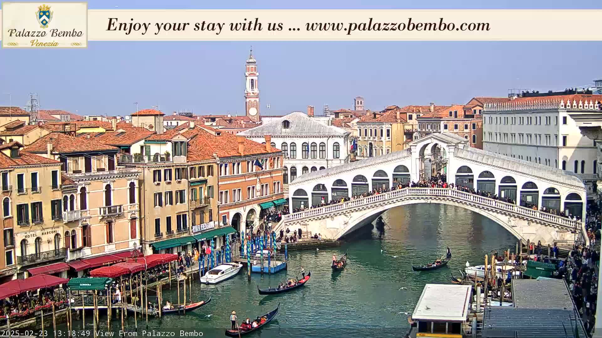 A sunny day in Venice shows a canal with gondolas and a bridge crowded with people, with buildings lining the waterway.