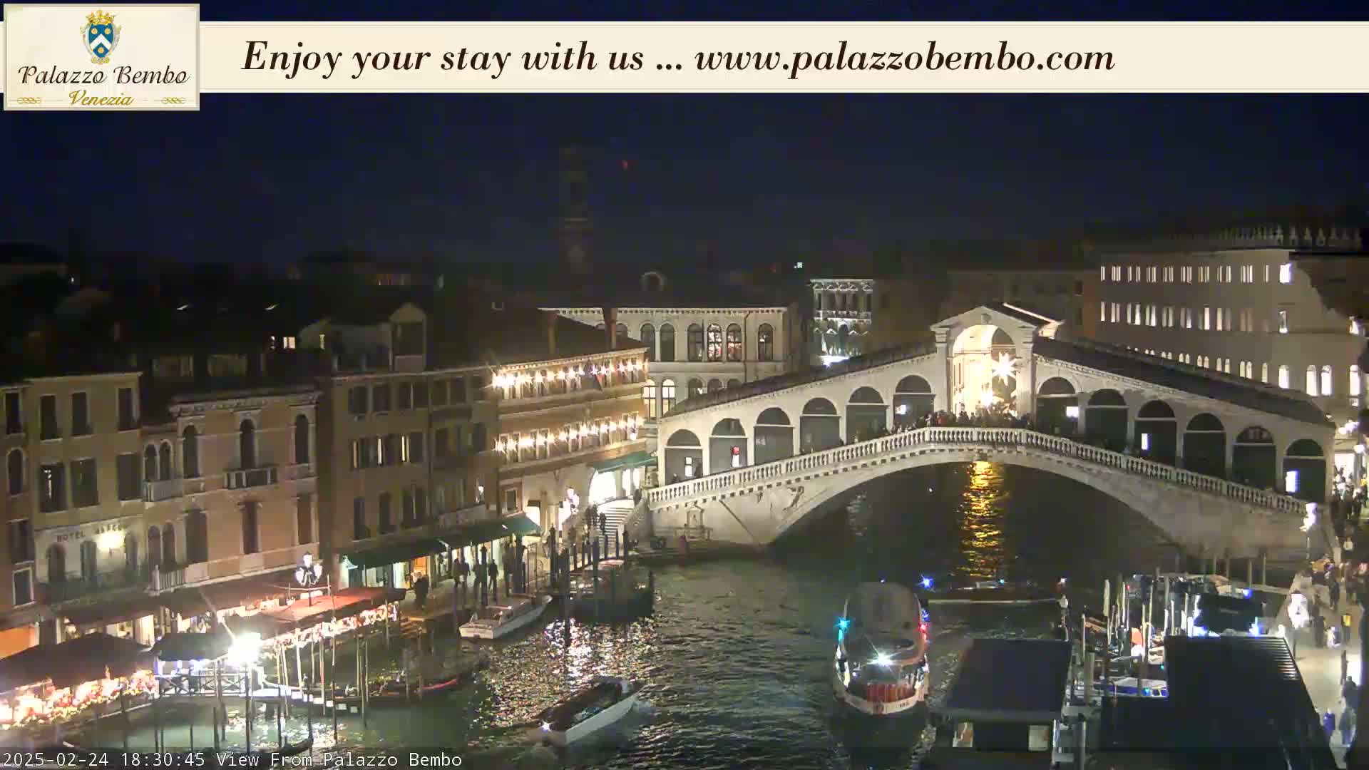 A nighttime view of a bridge over a canal in Venice, with illuminated buildings and boats lining the waterway.
