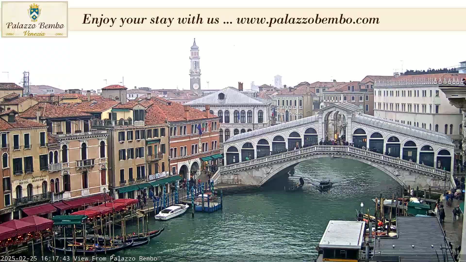 A view of a canal in Venice, Italy, showing a bridge with people walking across it, gondolas in the water, and buildings lining the canal on an overcast day.
