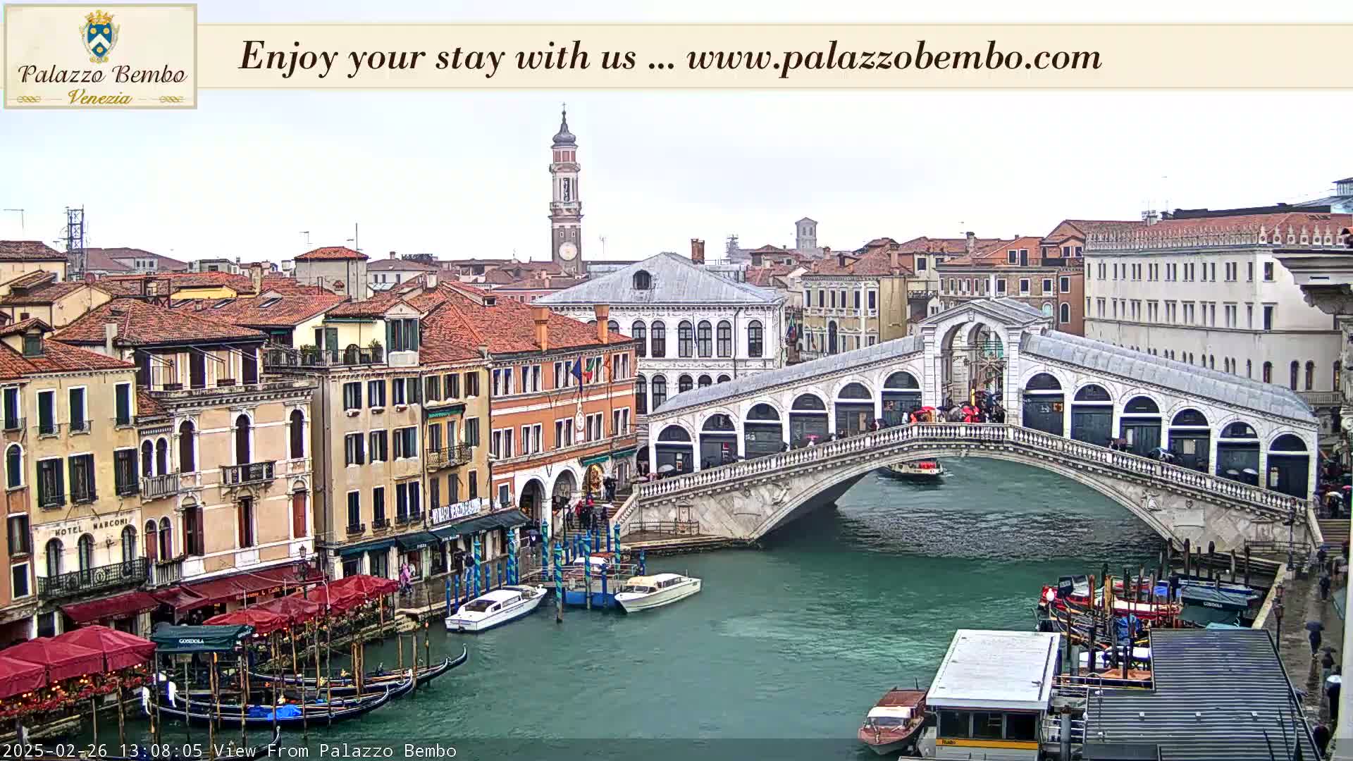 A view of a canal in Venice on an overcast day shows a bridge with pedestrians and gondolas, with buildings lining the waterway.