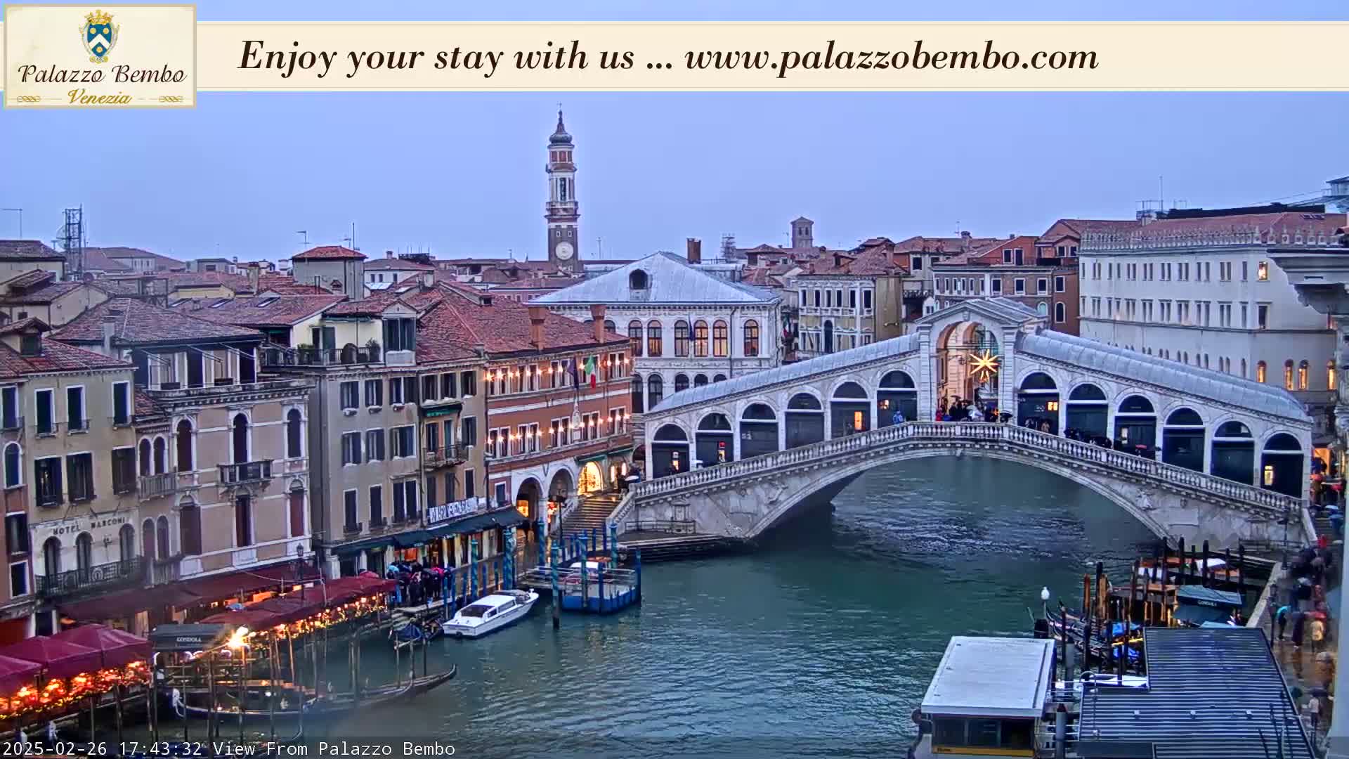 A twilight view of a canal in Venice, showing a bridge with pedestrians and boats, with buildings lining the waterway.