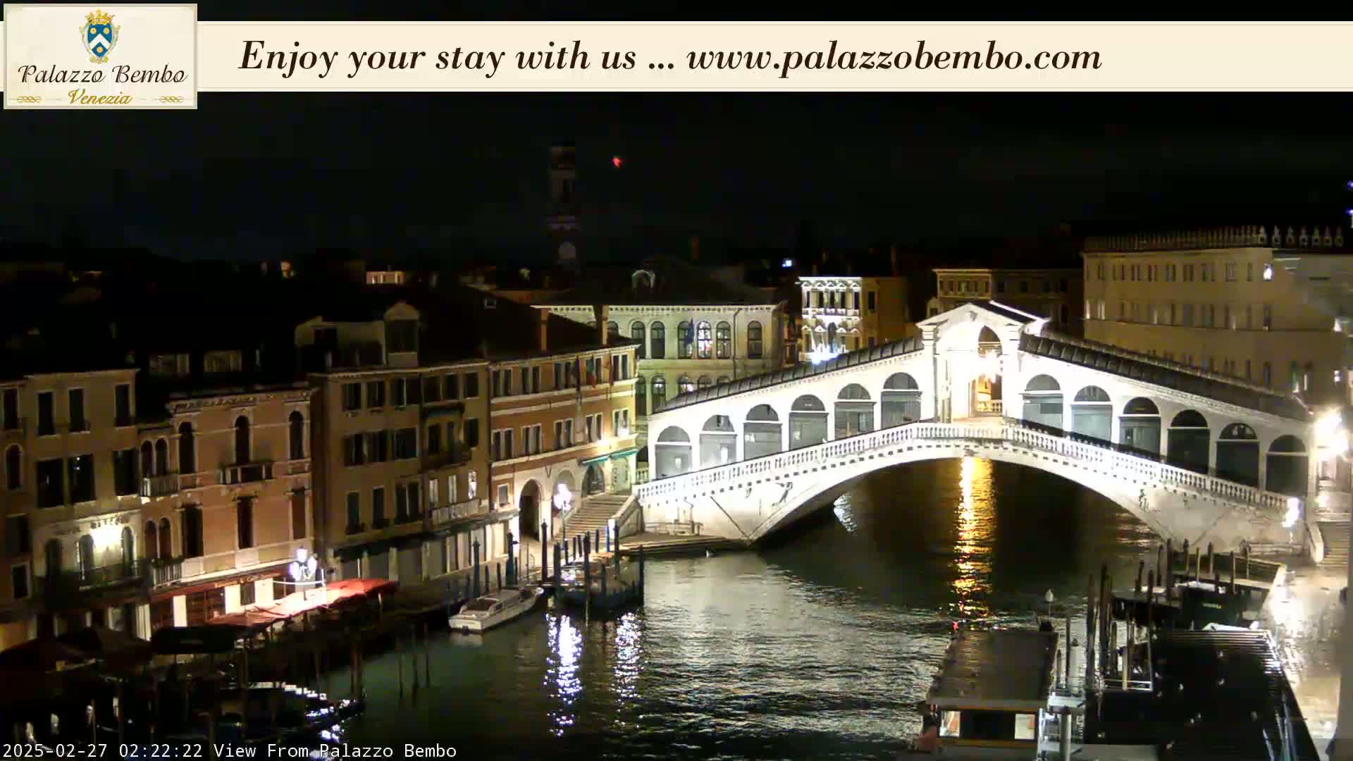 A nighttime view of a canal in Venice, showing a bridge illuminated against a dark sky.