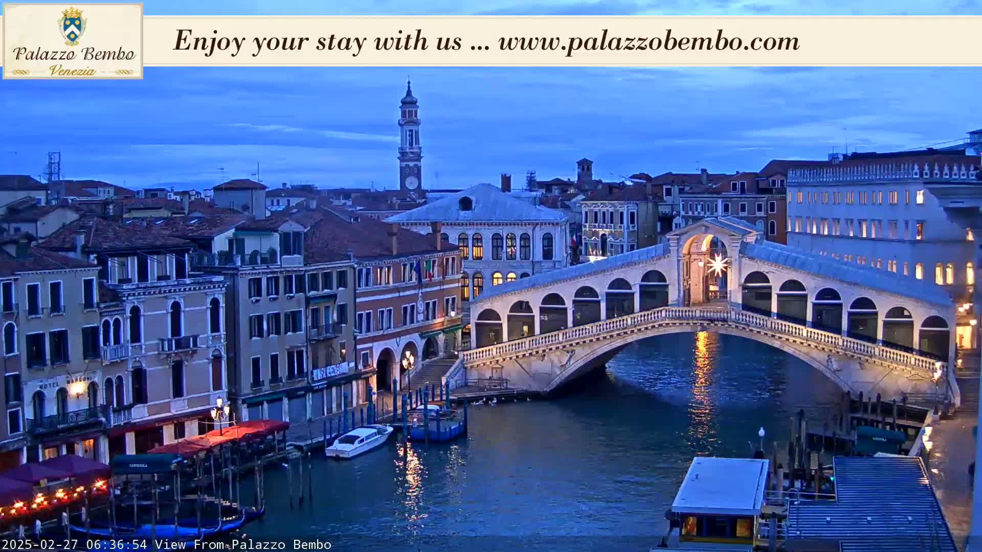 A twilight view of a canal in Venice, showing a bridge with arched walkways and buildings lining the waterway.