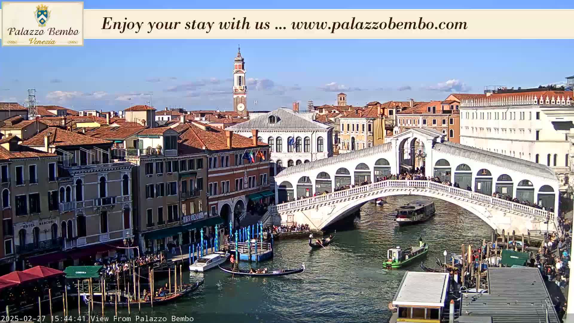 A sunny view of a Venetian canal, featuring gondolas, a bridge crowded with people, and numerous buildings along the waterway.