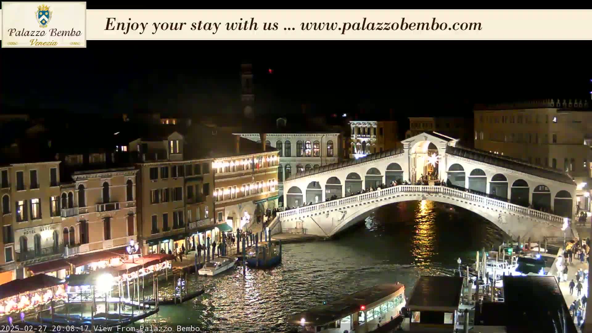 A brightly lit bridge spans a canal at night in Venice, with buildings and boats lining the waterway.