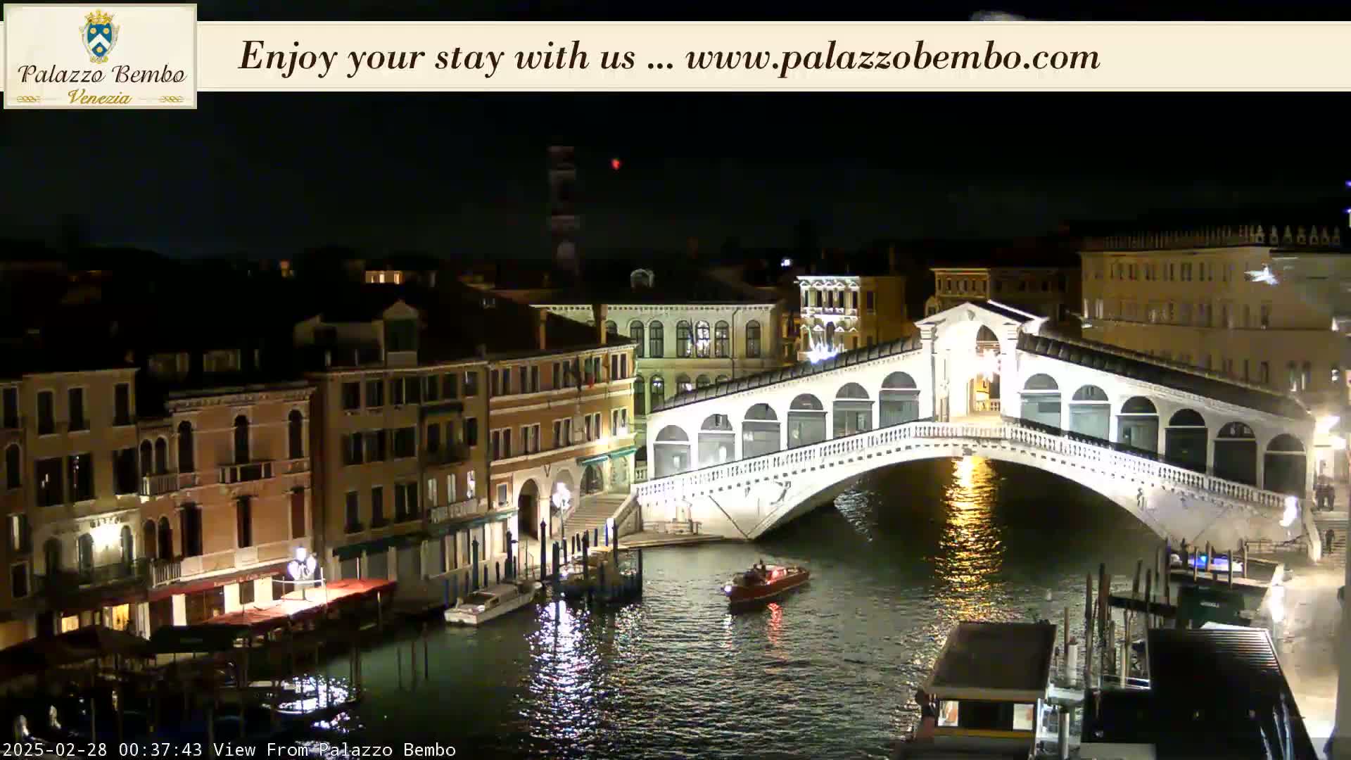 A nighttime view of a bridge spanning a canal in Venice, with boats and buildings illuminated along the waterway.