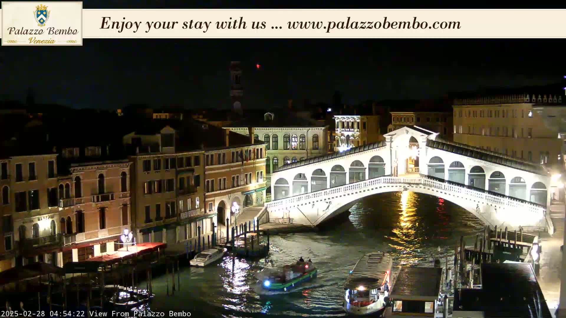 A nighttime view of a canal in Venice, showing the Rialto Bridge illuminated, with boats on the water and buildings lining the canal.