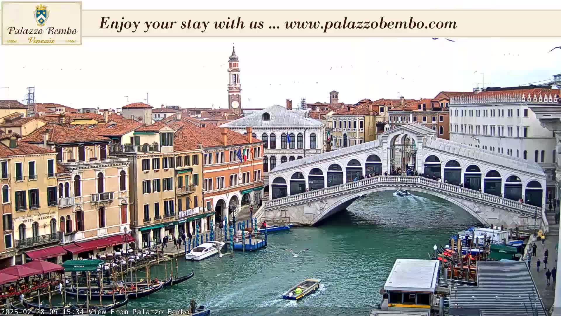 A view of a canal in Venice, Italy, shows a bridge with people walking across it, boats on the water, and buildings lining the canal under an overcast sky.