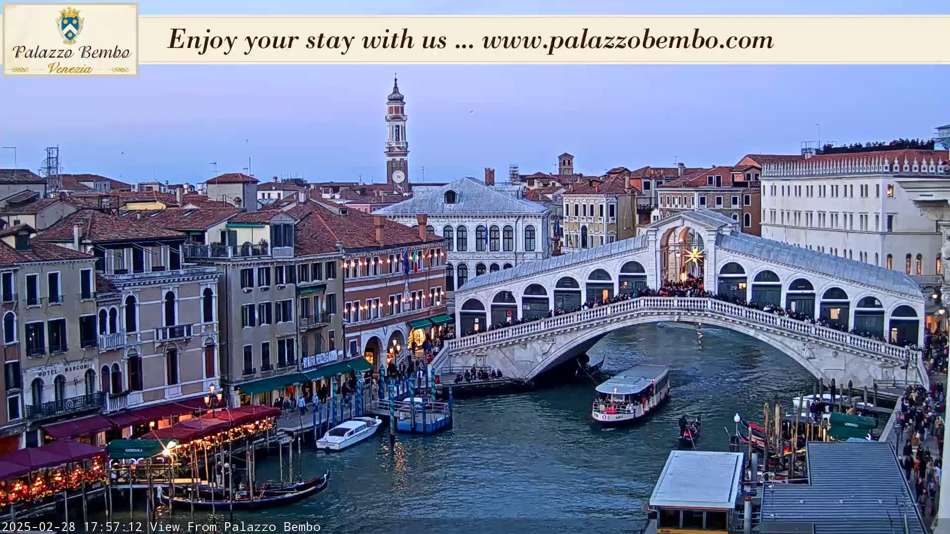 A twilight view of the Rialto Bridge in Venice, Italy, with numerous people on the bridge and gondolas and boats in the canal below.