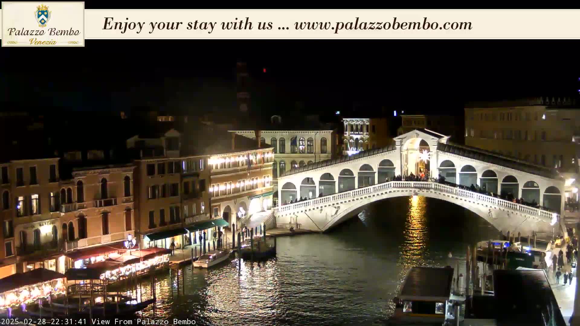 A nighttime view of a bridge spanning a canal in Venice, with illuminated buildings lining the waterway.