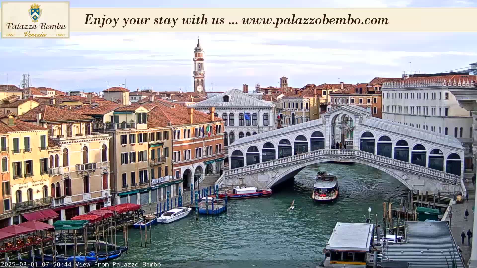 A canal in Venice, under a partly cloudy sky, shows a bridge with pedestrians and boats passing underneath, and numerous buildings lining the waterway.