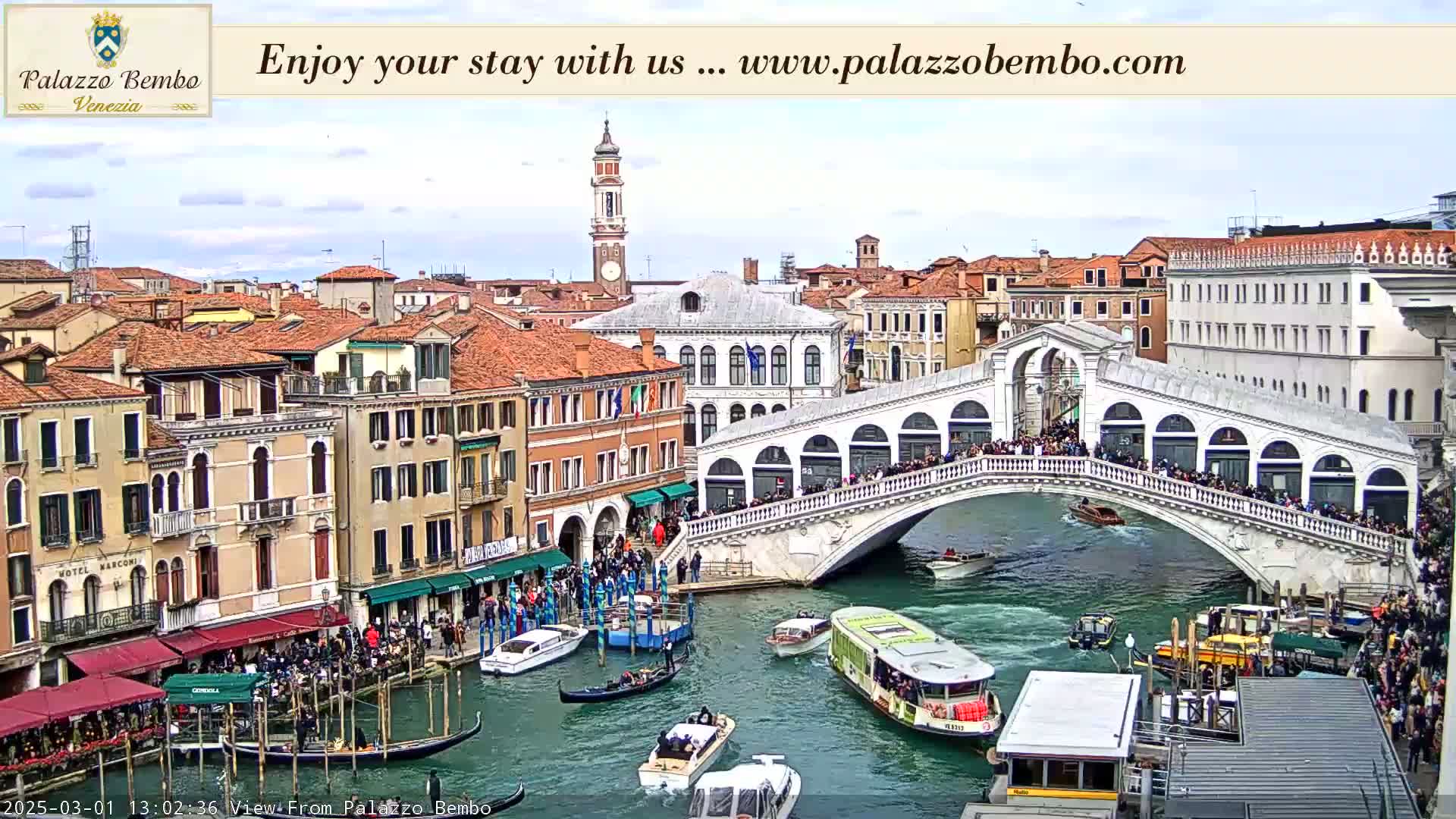 A crowded Venetian bridge arches over a canal filled with gondolas and water taxis under a partly cloudy sky.