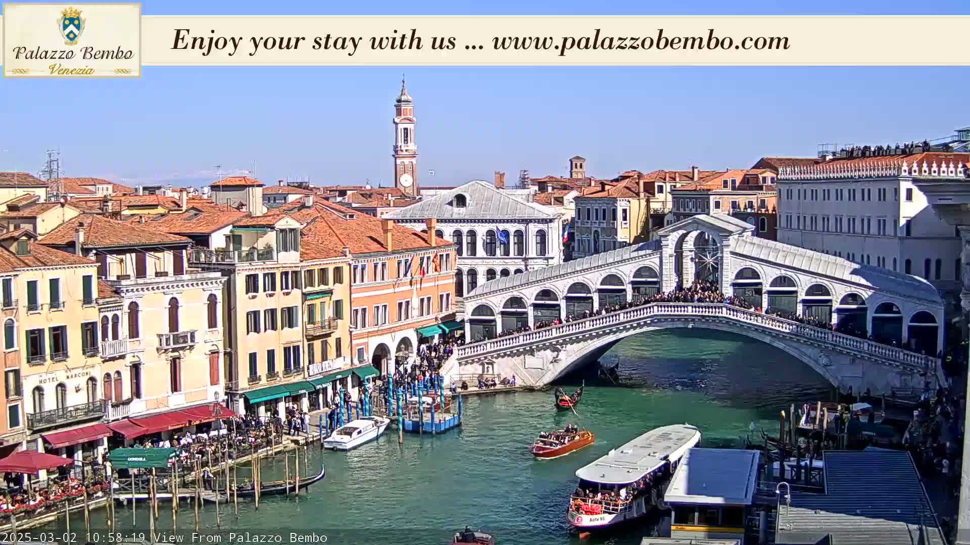 A sunny view of a Venetian canal with gondolas and a crowded bridge, surrounded by historic buildings.