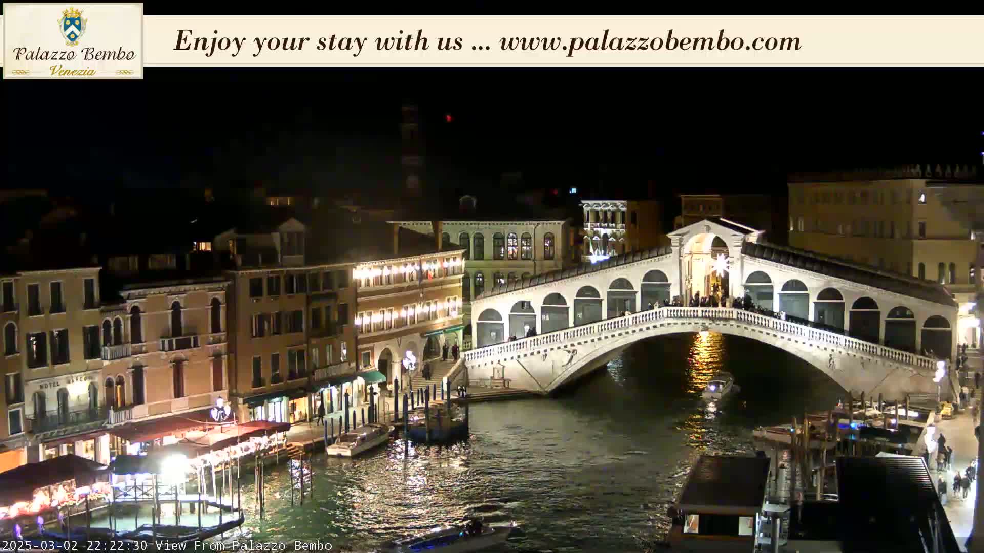 A nighttime view of a bridge spanning a canal in Venice, with illuminated buildings lining the waterways.
