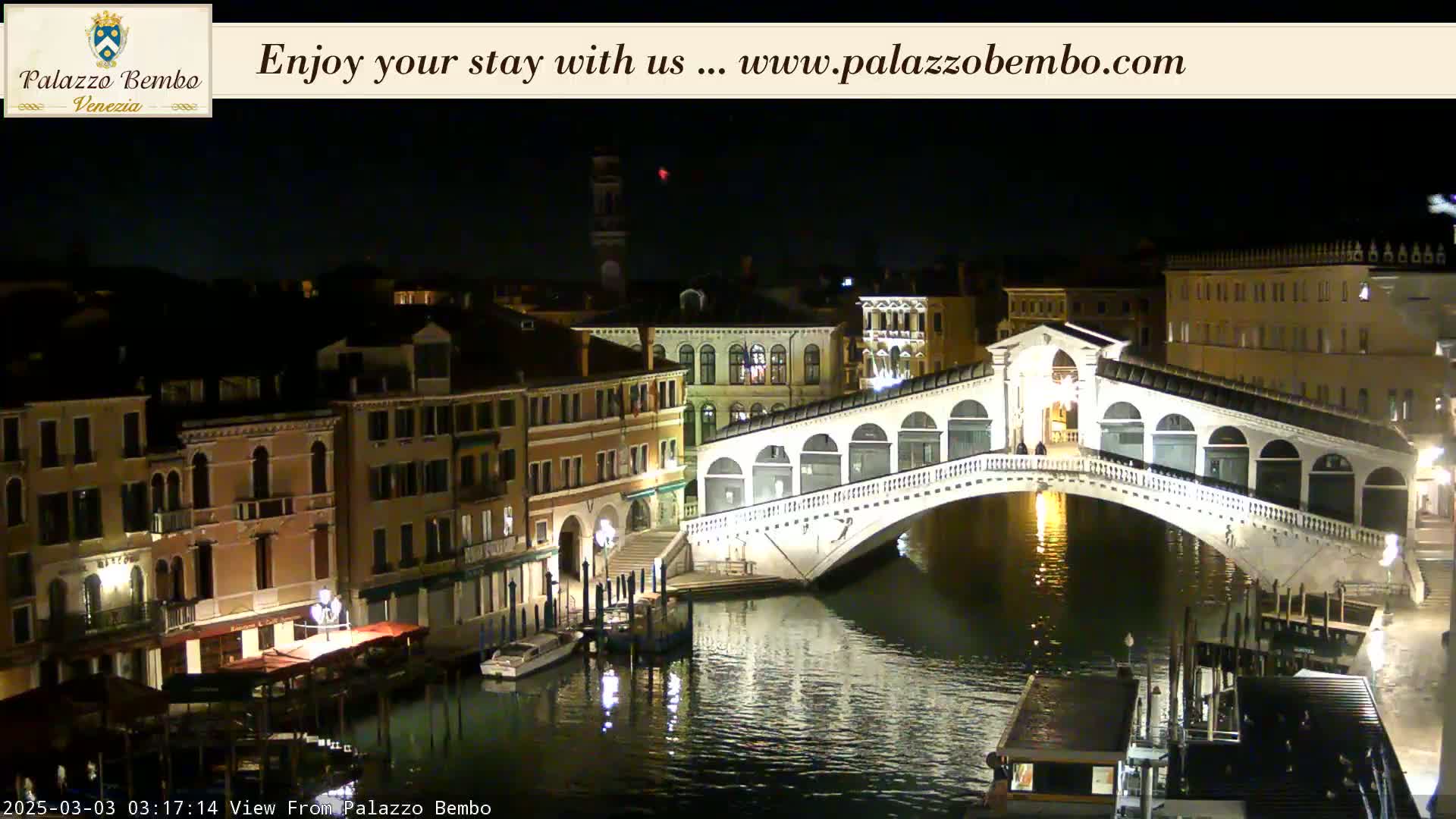 A nighttime view of a canal in Venice, showing a bridge lit up and buildings lining the waterway.
