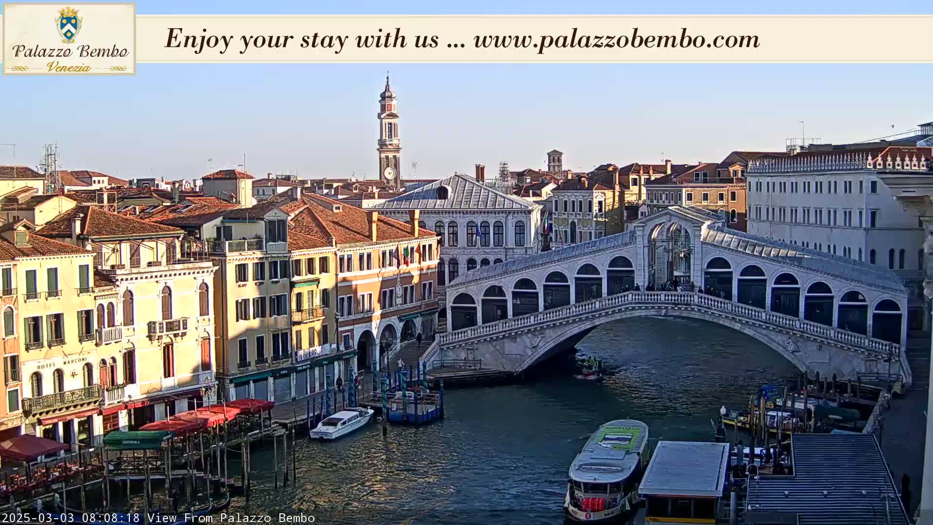 A sunny view of a canal in Venice, Italy, shows a bridge with pedestrians crossing it, boats on the water, and colorful buildings lining the canal.
