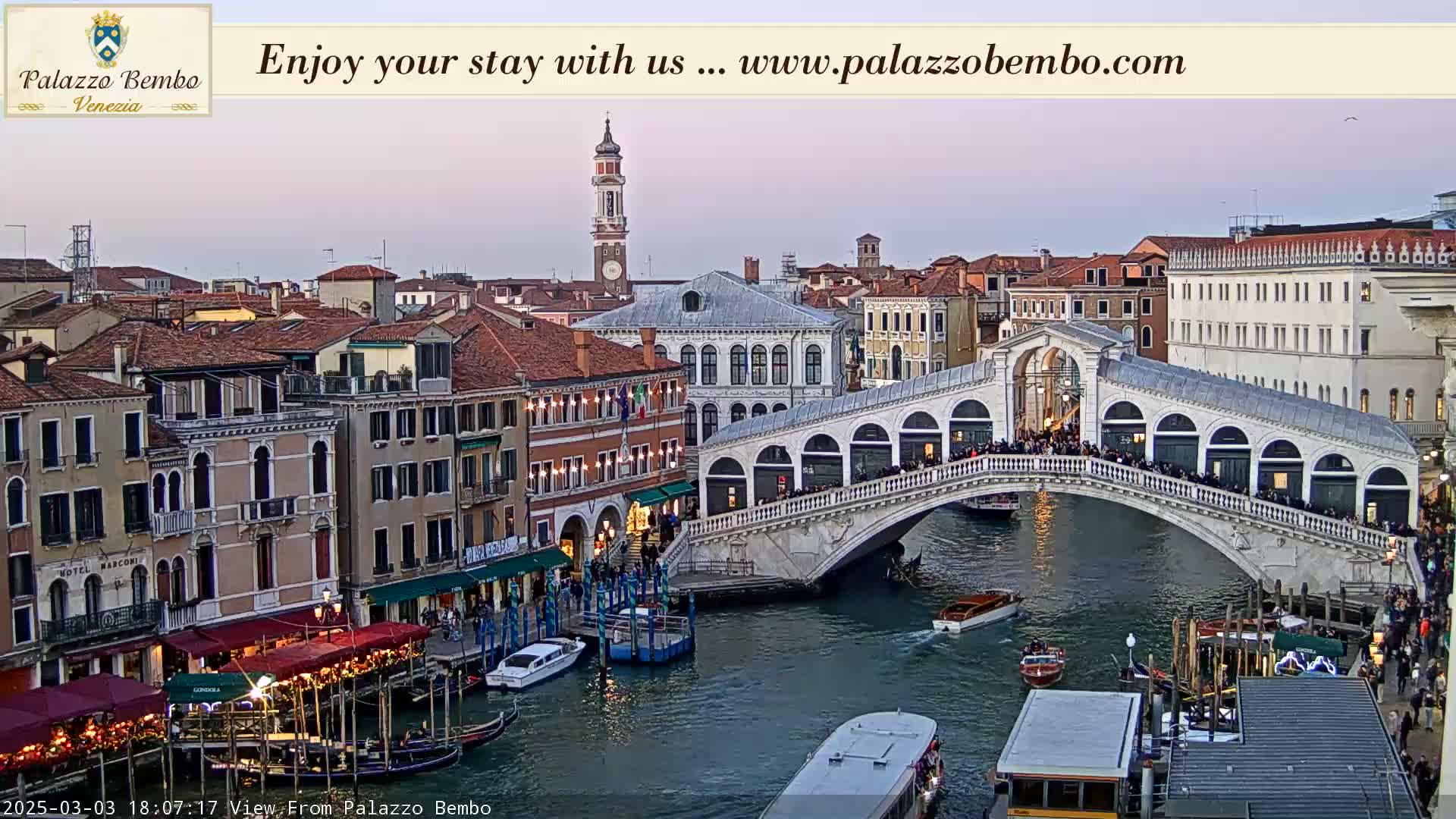 A bridge over a canal in Venice is crowded with people at dusk, with boats and buildings lining the waterway.