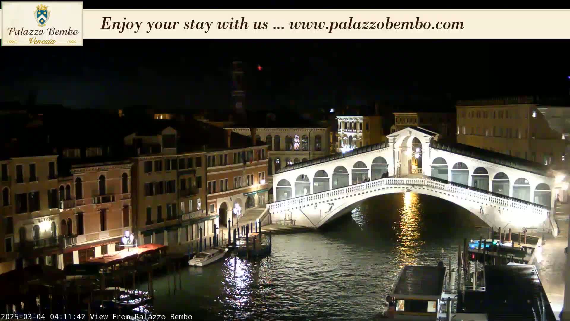 A nighttime view of a canal in Venice, showing a white bridge illuminated against a dark sky.