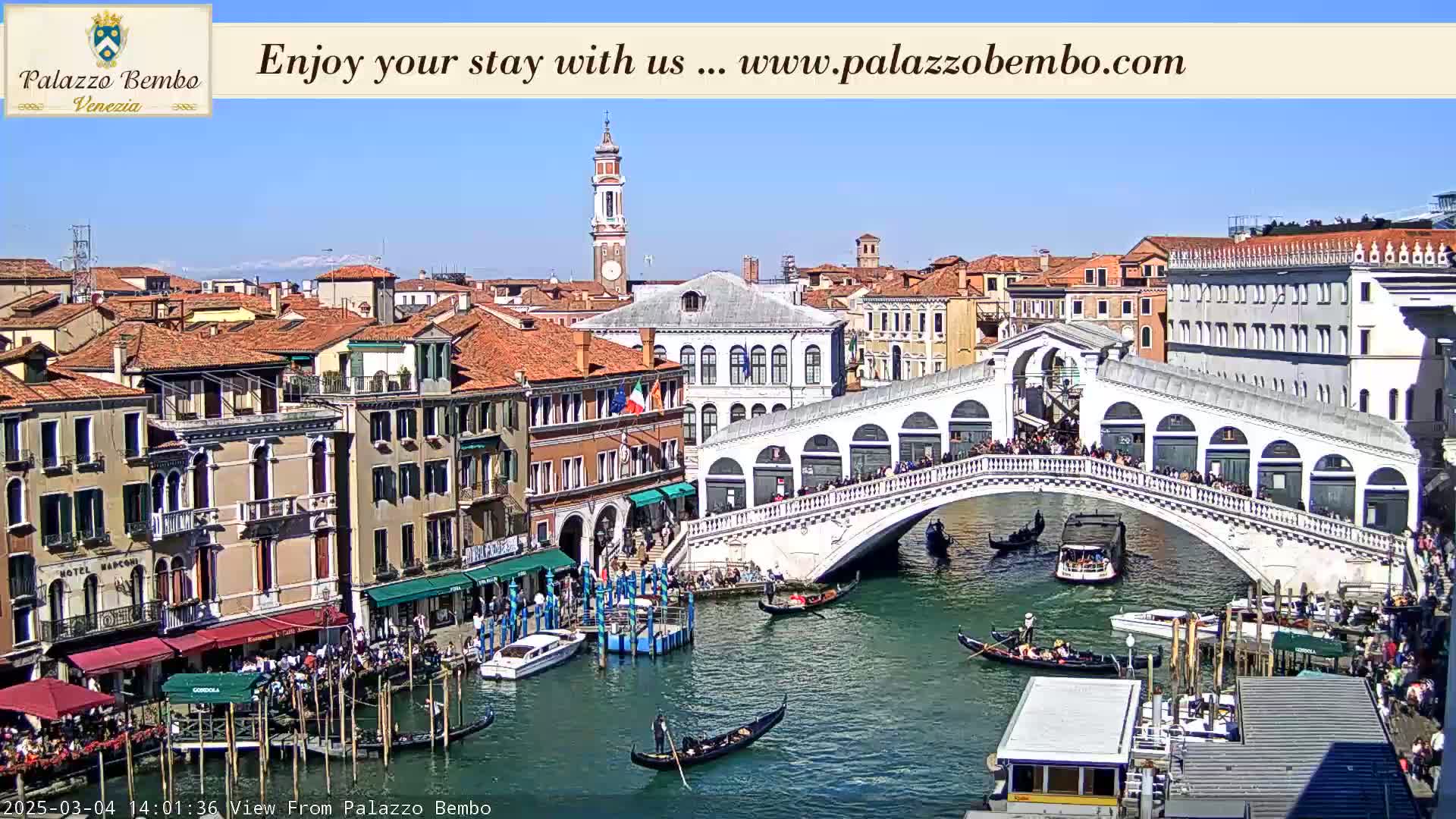 A sunny view of a Venetian canal with gondolas, a bridge crowded with people, and colorful buildings lining the waterway.