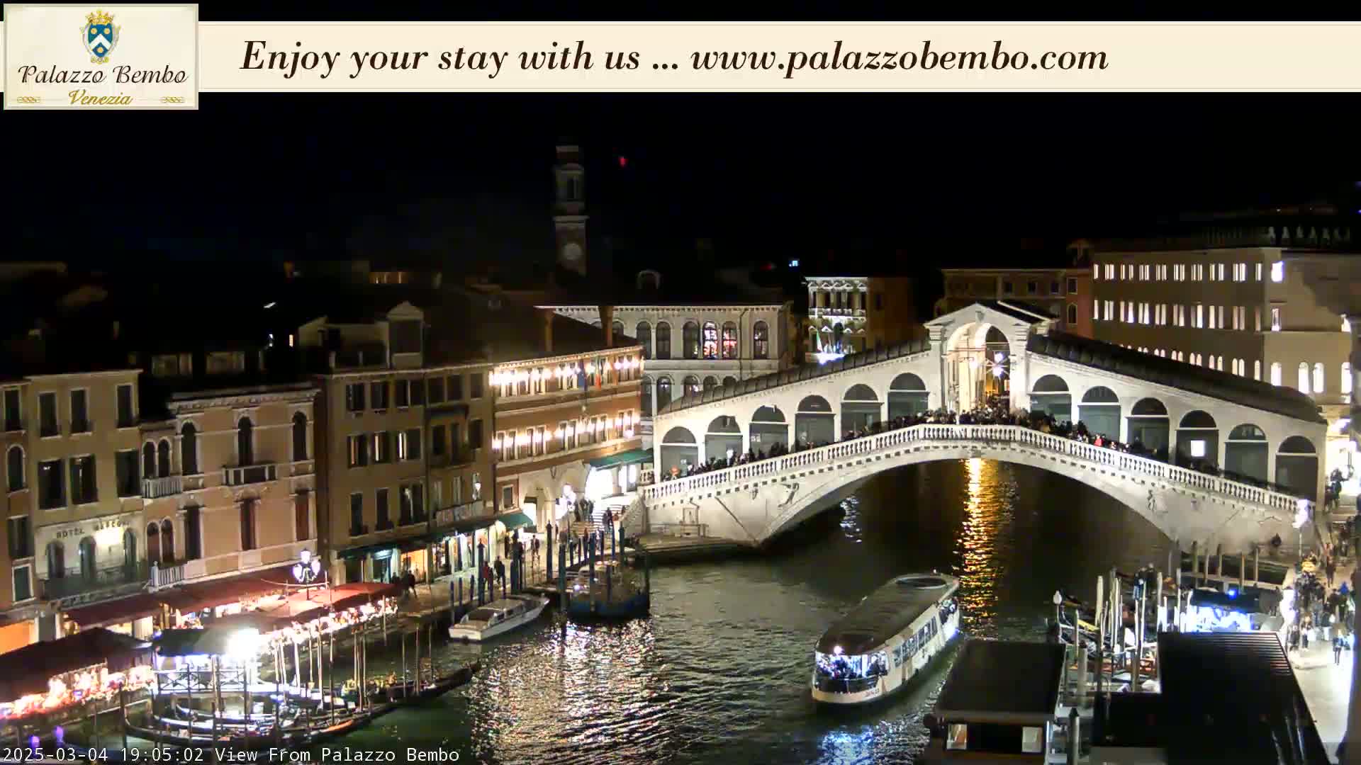 A nighttime view of a bridge over a canal in Venice, with many people on the bridge and boats in the water, under a clear, dark sky.