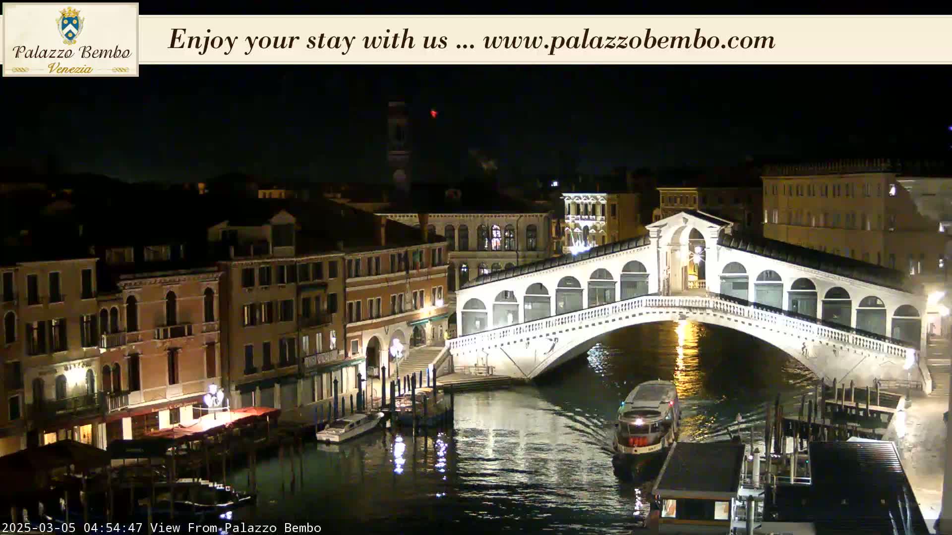 A nighttime view of a canal in Venice, showing a bridge with boats and buildings illuminated along the waterway under a clear, dark sky.
