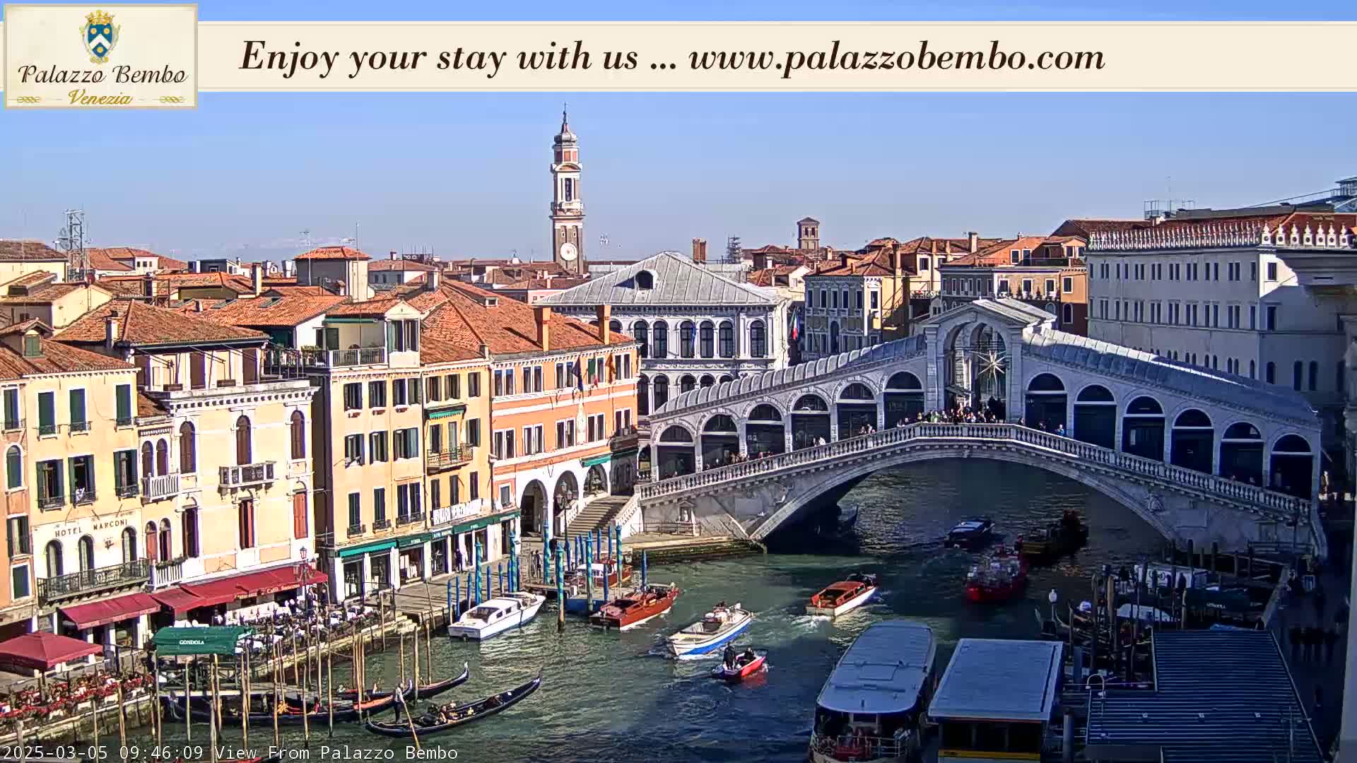A sunny view of a canal in Venice, Italy, shows a bridge crowded with people, boats on the water, and numerous buildings lining the waterway.