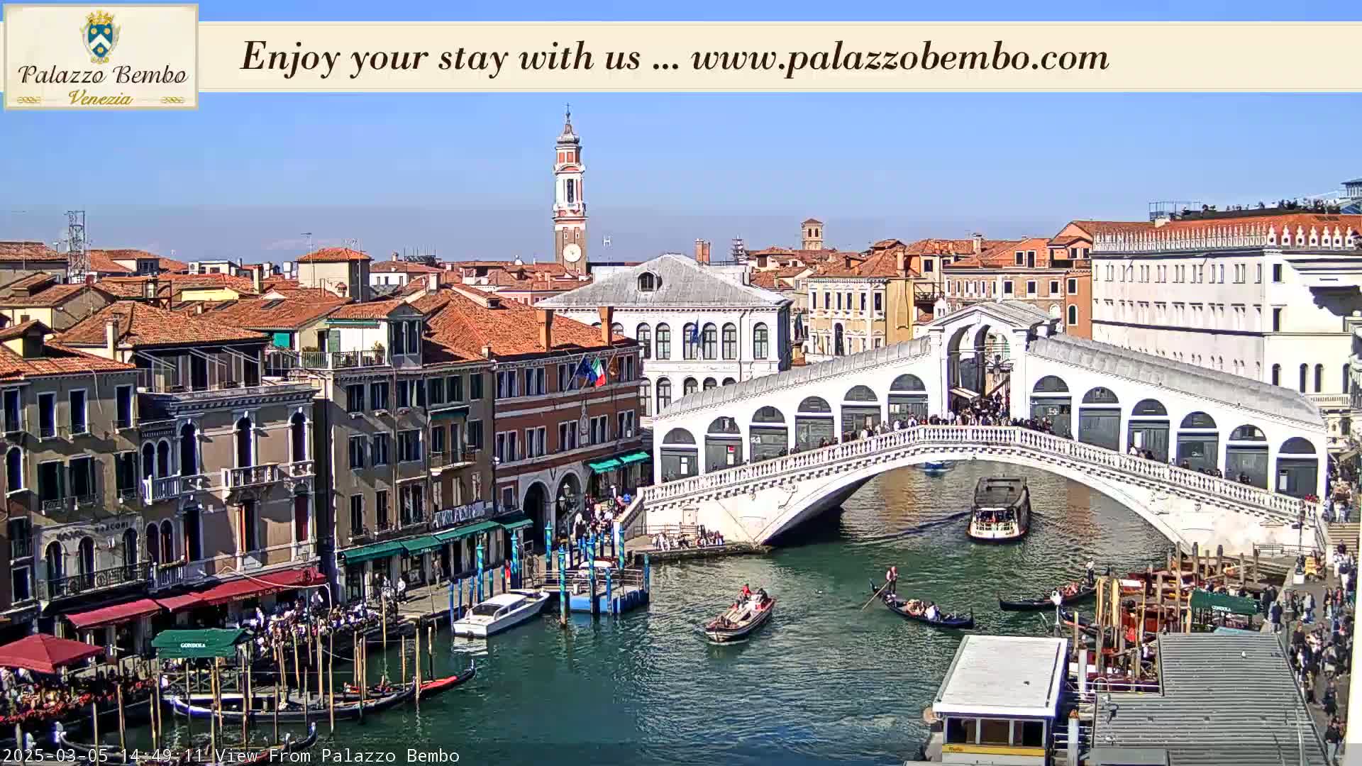 A sunny day in Venice shows a canal with gondolas, boats, and a bridge crowded with people, all surrounded by historic buildings.