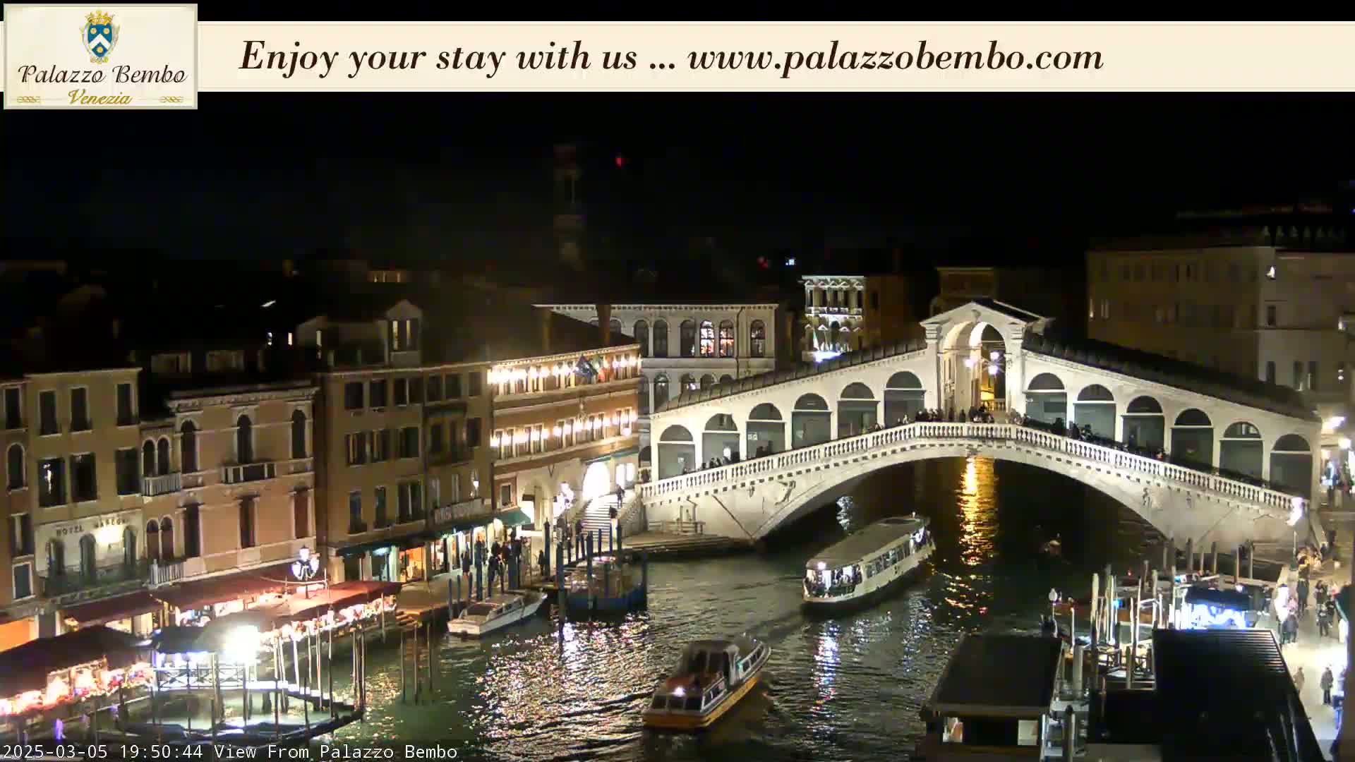 At night, a bridge stretches over a canal in Venice, with boats and illuminated buildings lining the waterways.