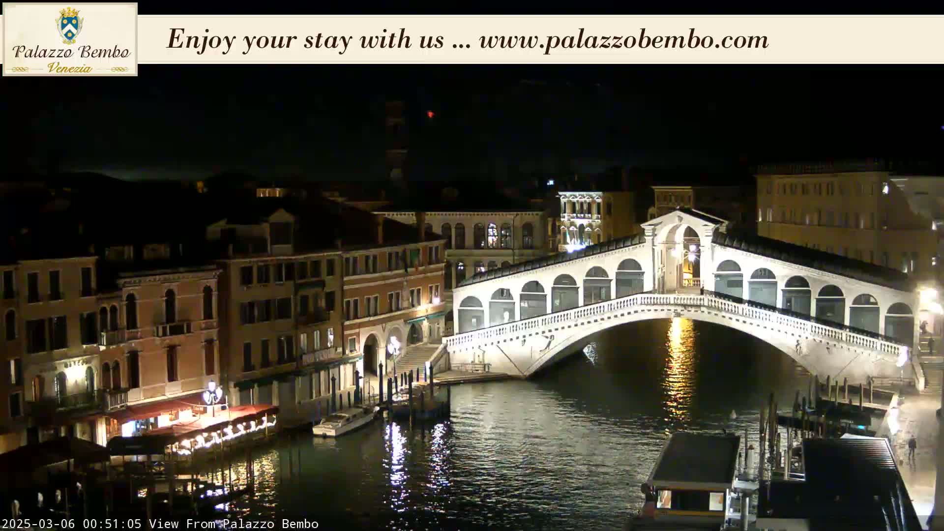 A nighttime view of a canal in Venice, showing a lit bridge and buildings along the waterway under a dark sky.