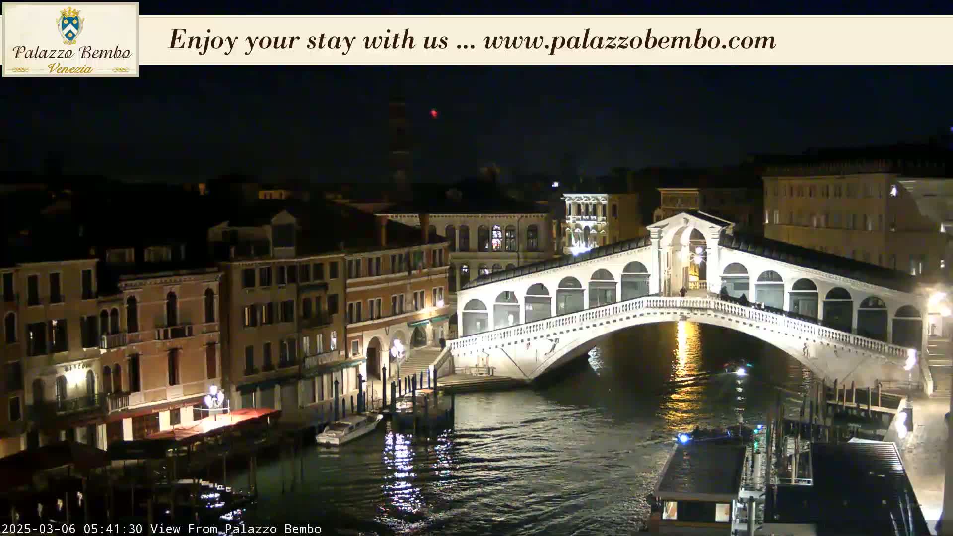A nighttime view of a canal in Venice, showing the Rialto Bridge lit up and boats on the water.