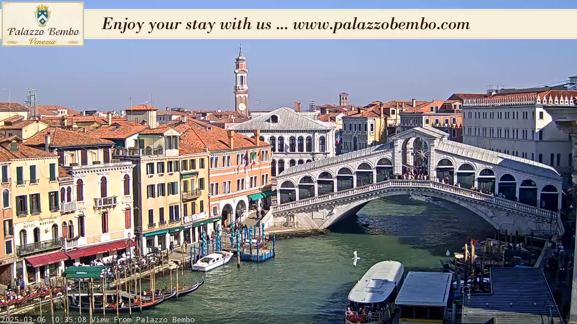 A sunny view of a canal in Venice, showing gondolas, a bridge with people on it, and buildings lining the waterway.
