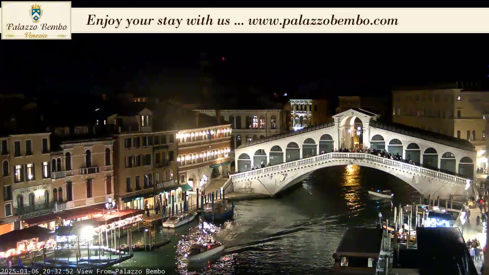 A nighttime view of a canal in Venice, showing a lit bridge with people on it, buildings along the canal's edge, and boats on the water.