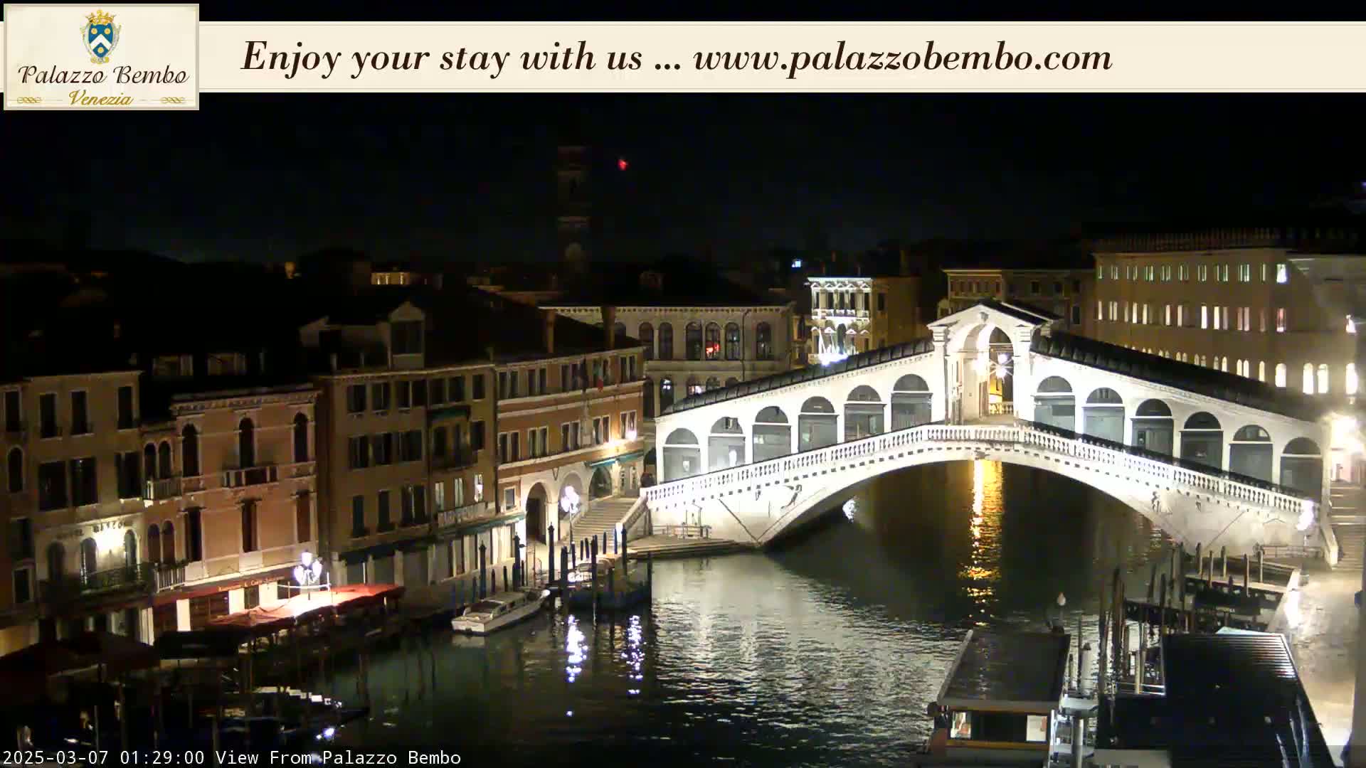A nighttime view of a canal in Venice, showing a bridge lit up against a dark sky.