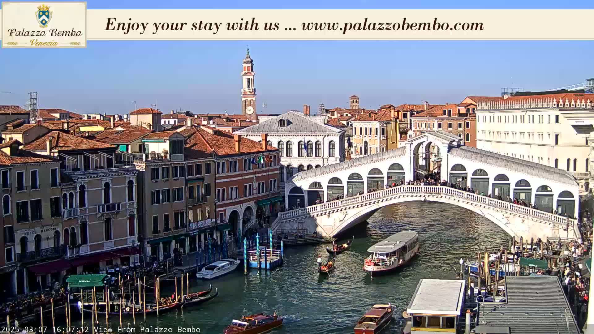 A sunny day in Venice shows the Rialto Bridge with people walking across it and gondolas and water taxis navigating the canal below, surrounded by historic buildings.
