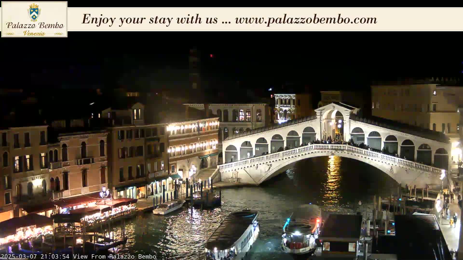 A nighttime view of a bridge over a canal in Venice, with illuminated buildings lining the waterway and boats moored below.