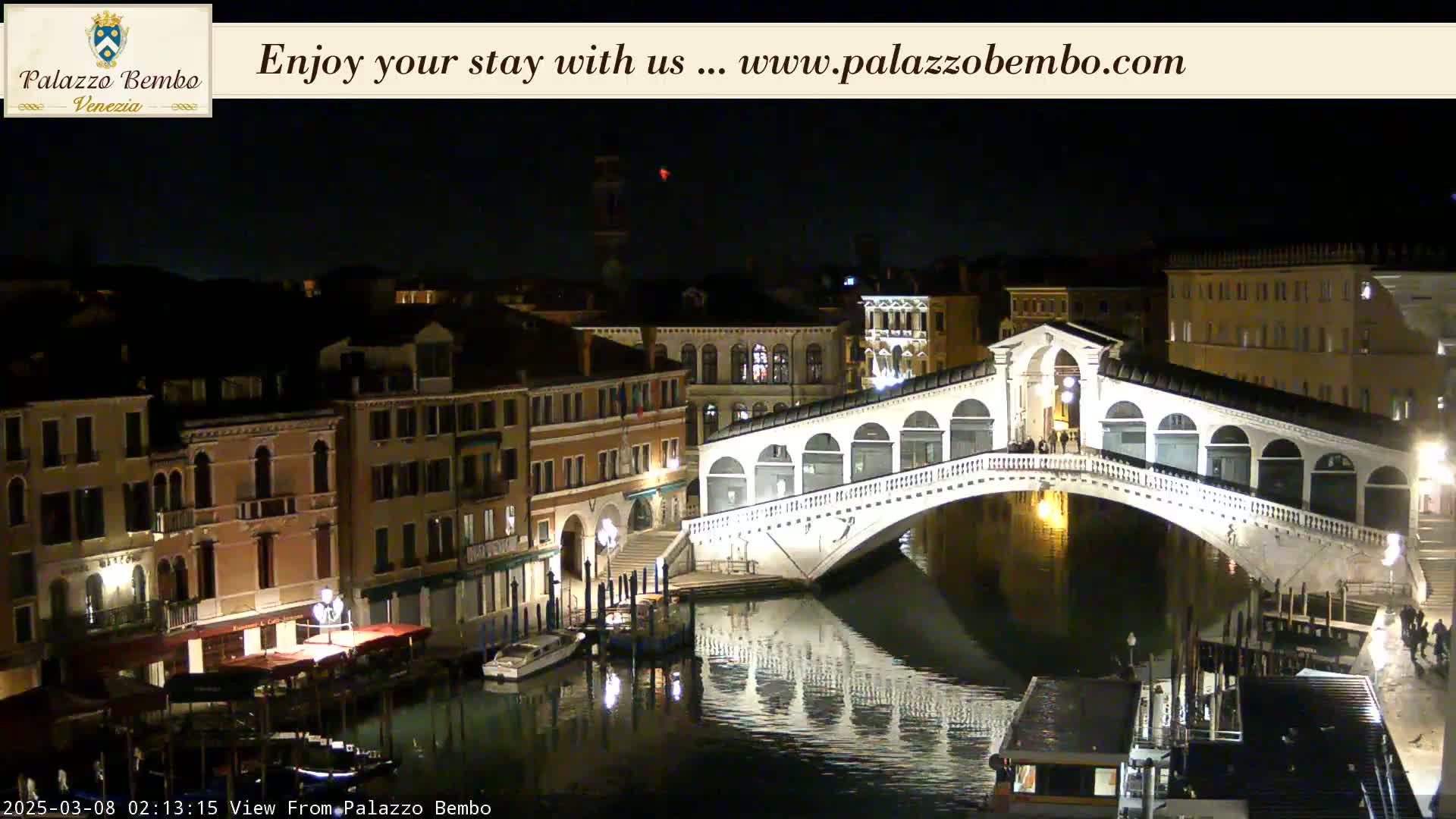 A nighttime view of a canal in Venice, showing the Rialto Bridge lit up and reflecting in the water, with buildings lining the waterway.