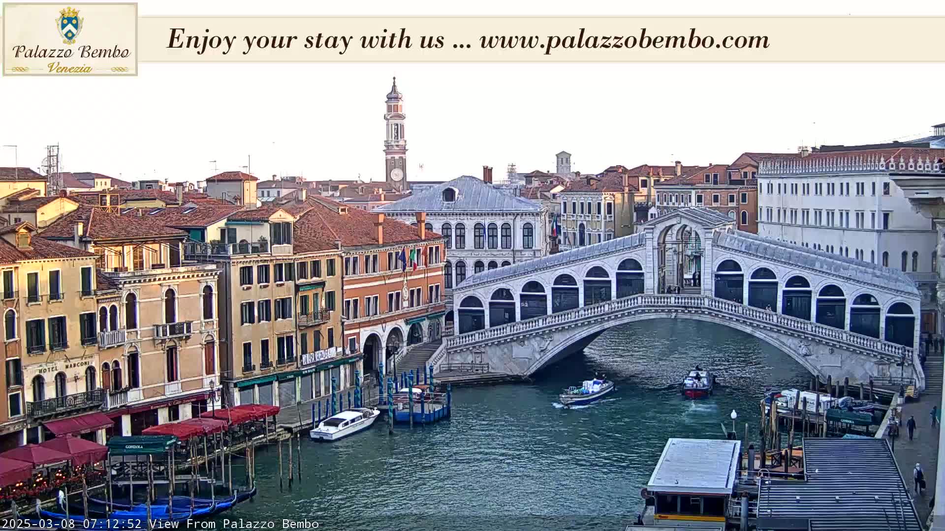 A Venetian canal scene shows a bridge arching over the water, with colorful buildings lining the waterway, boats moored along the banks, and a mostly clear sky.