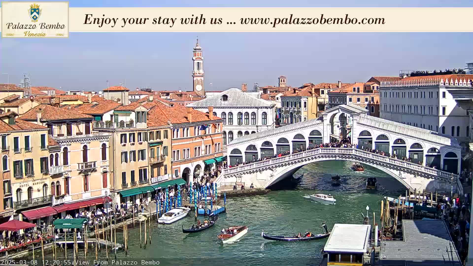 A sunny day in Venice shows the Rialto Bridge crowded with people, gondolas and other boats navigating the canal below, with many buildings lining the waterway.