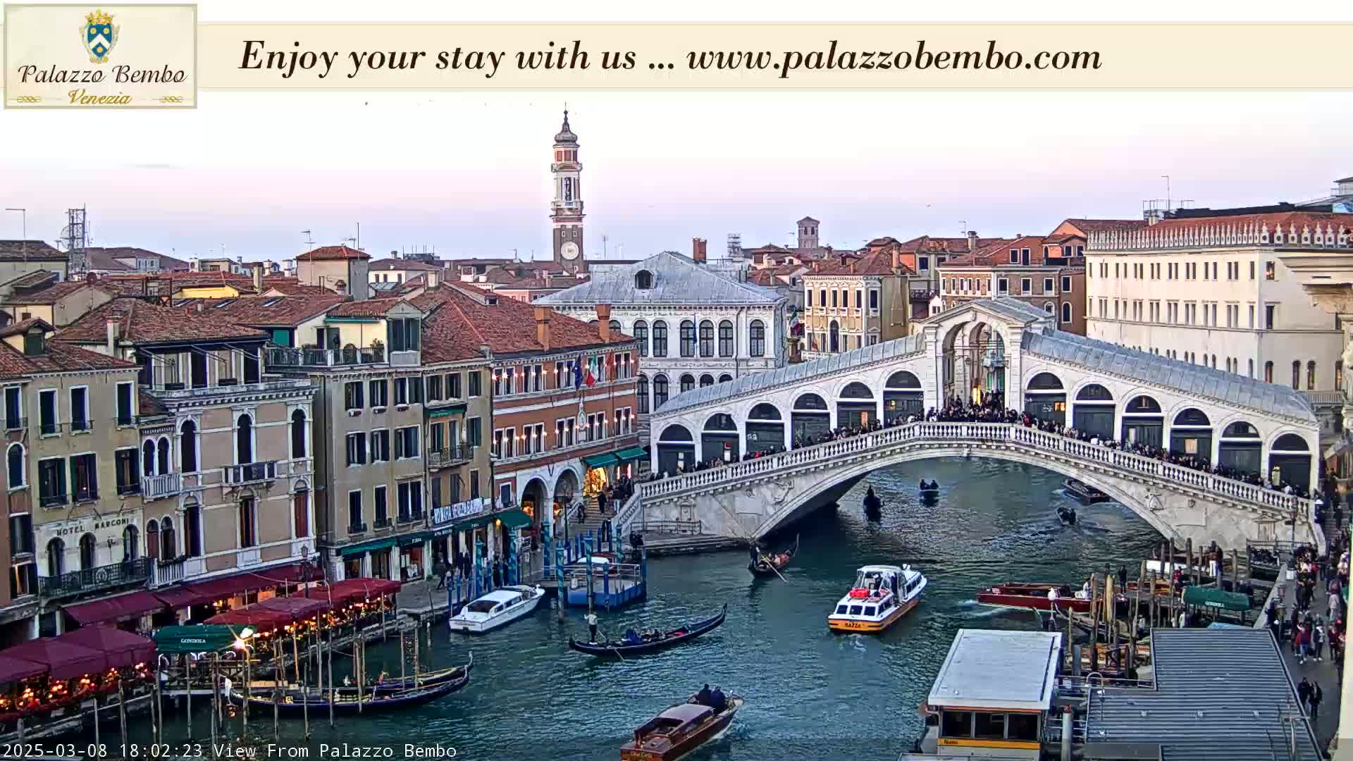 A twilight view of a Venetian bridge crowded with people, with gondolas and other boats on the canal below, and buildings lining the waterfront.