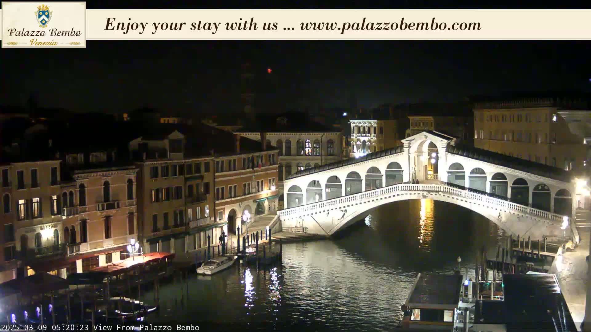 A nighttime view of a canal in Venice, showing a bridge illuminated with lights, boats, and buildings along the water's edge.