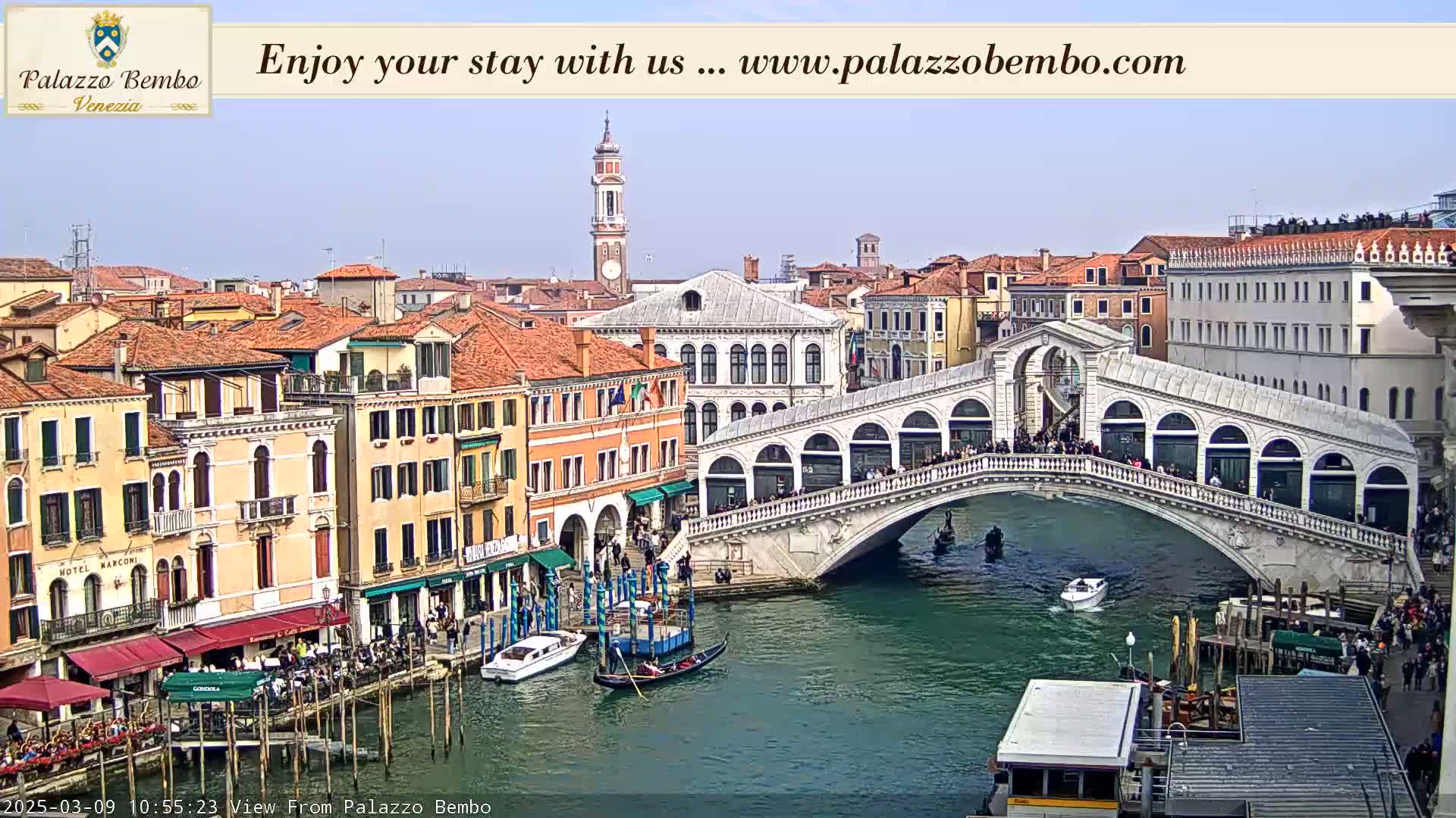 A view of a canal in Venice, Italy, on a partly sunny day shows the Rialto Bridge with pedestrians crossing it and gondolas and boats on the water below, along with buildings lining the canal.