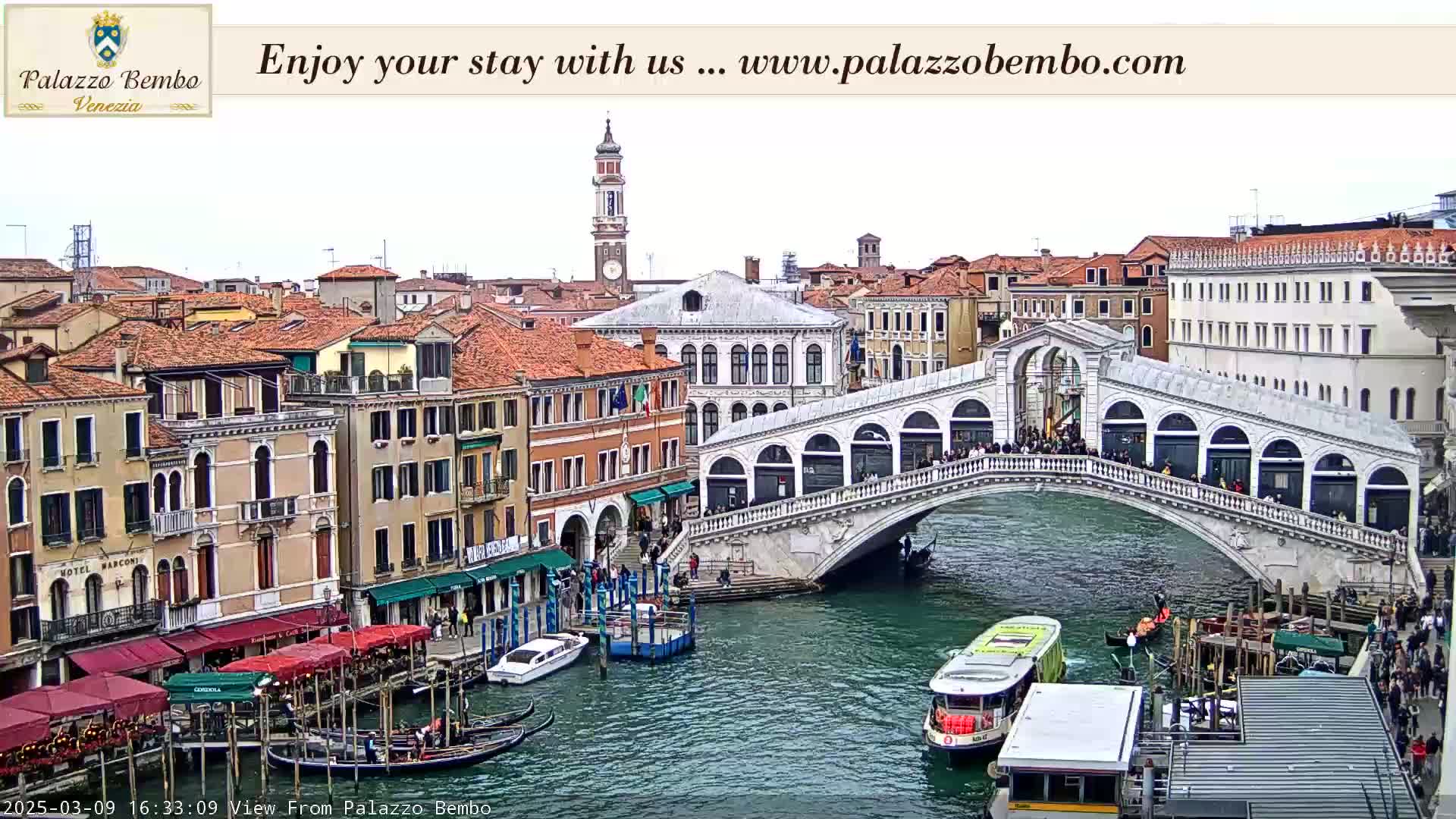 A view of a canal in Venice, showing a bridge with people walking across it and gondolas moored along the sides of the buildings under an overcast sky.