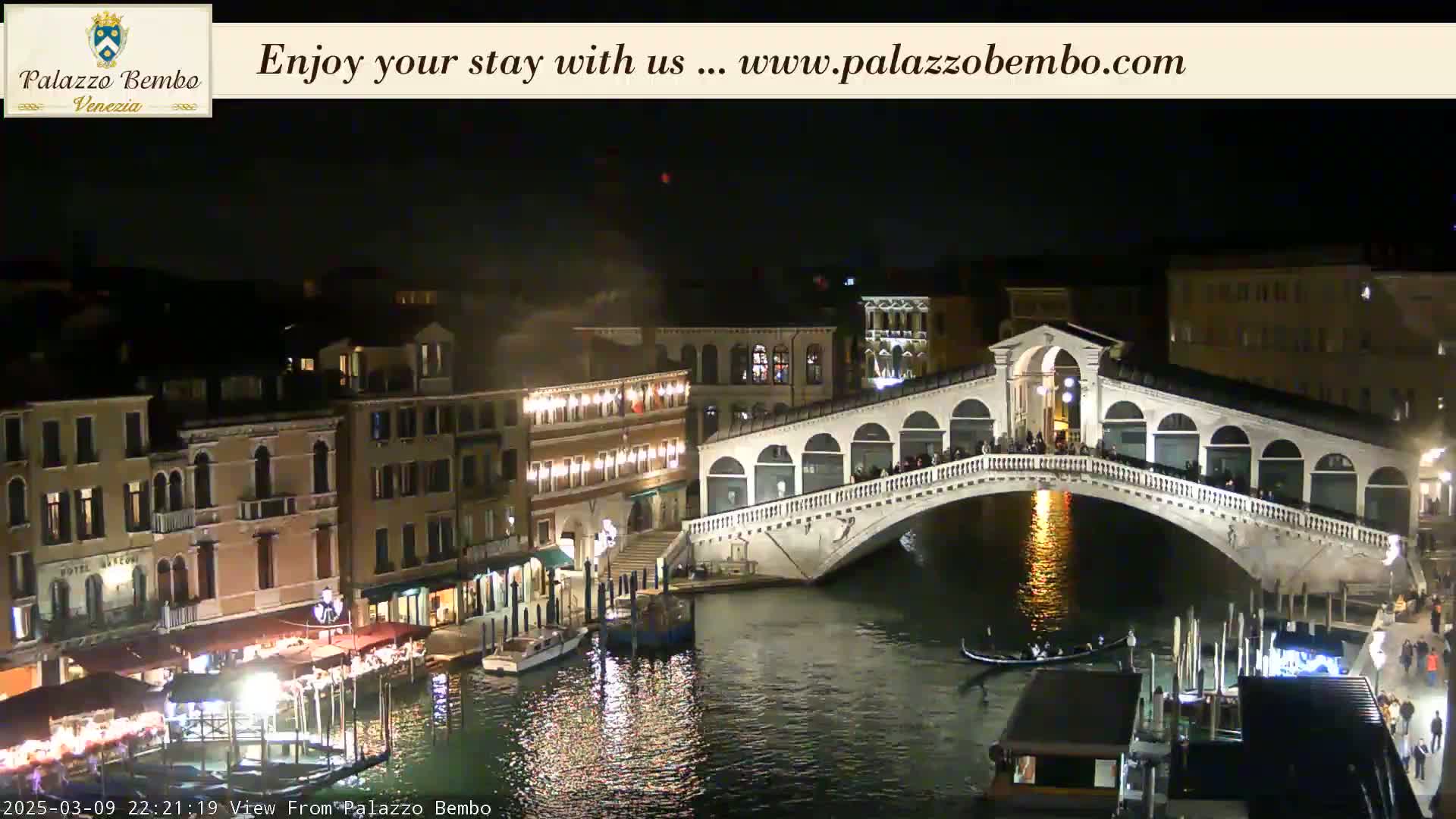 A nighttime view of a bridge over a canal in Venice, with illuminated buildings lining the waterway and people walking across the bridge.