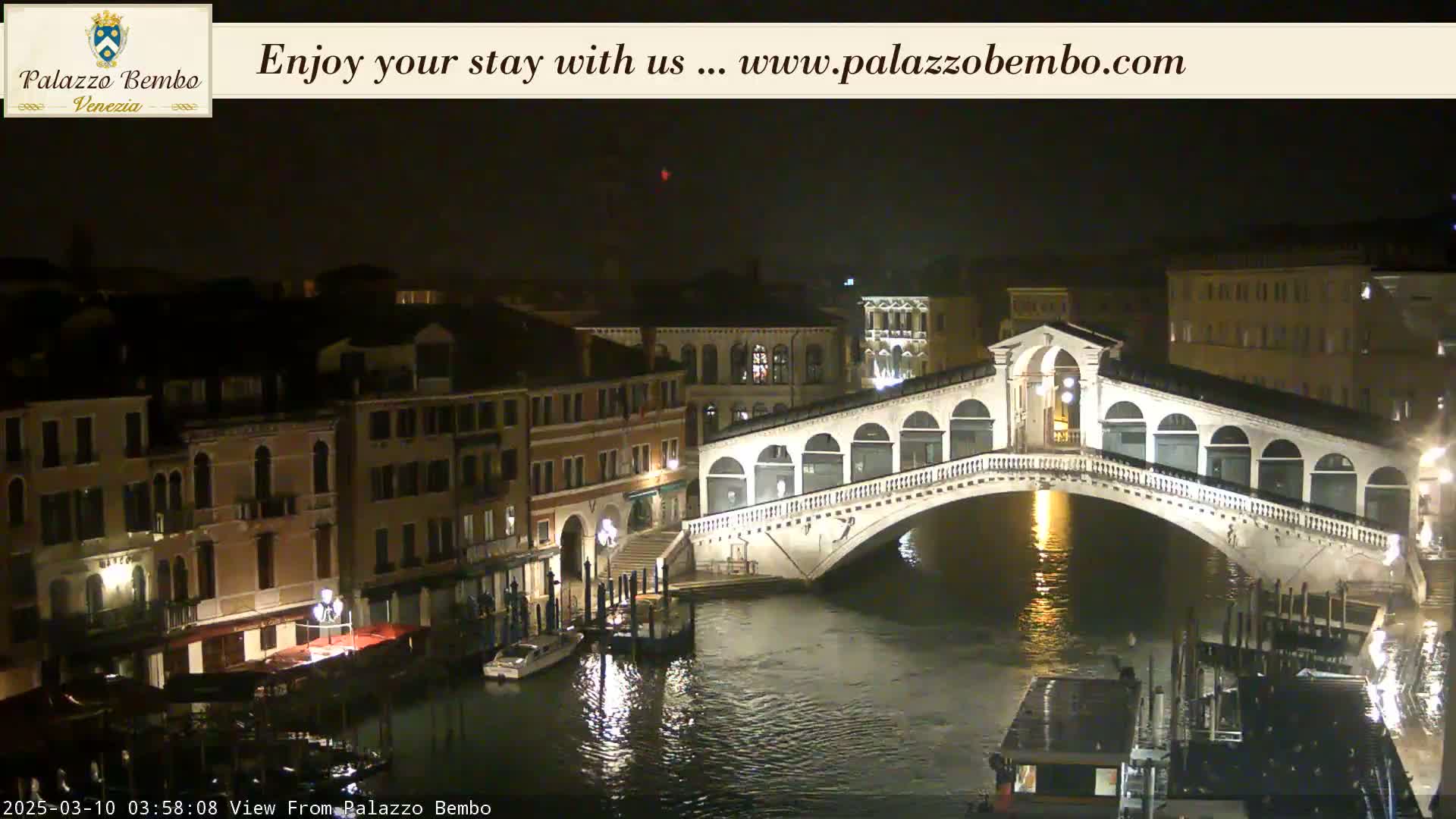 A nighttime view of a bridge over a canal in Venice, with buildings illuminated along the waterway under a dark sky.