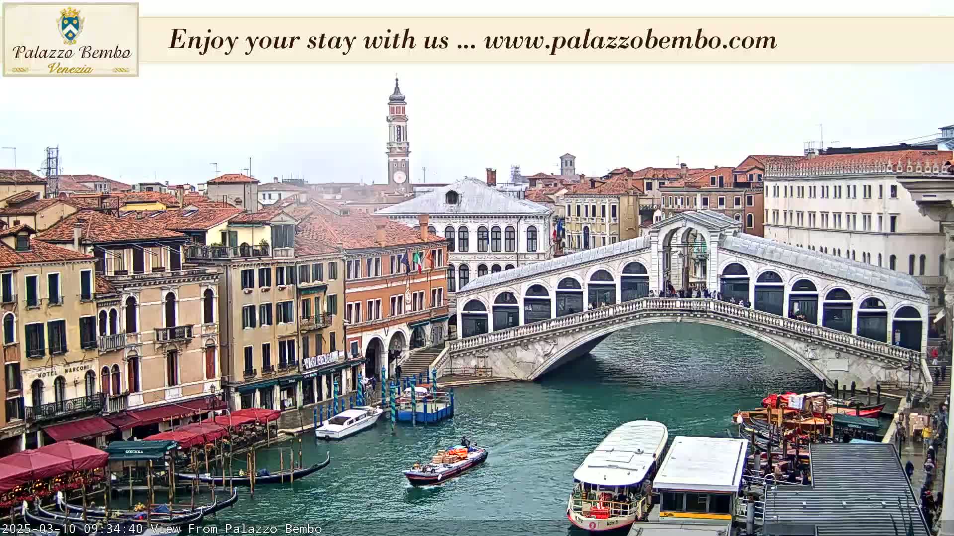 A canal in Venice, under an overcast sky, features a bridge with people on it, boats passing underneath, and buildings lining the waterway.