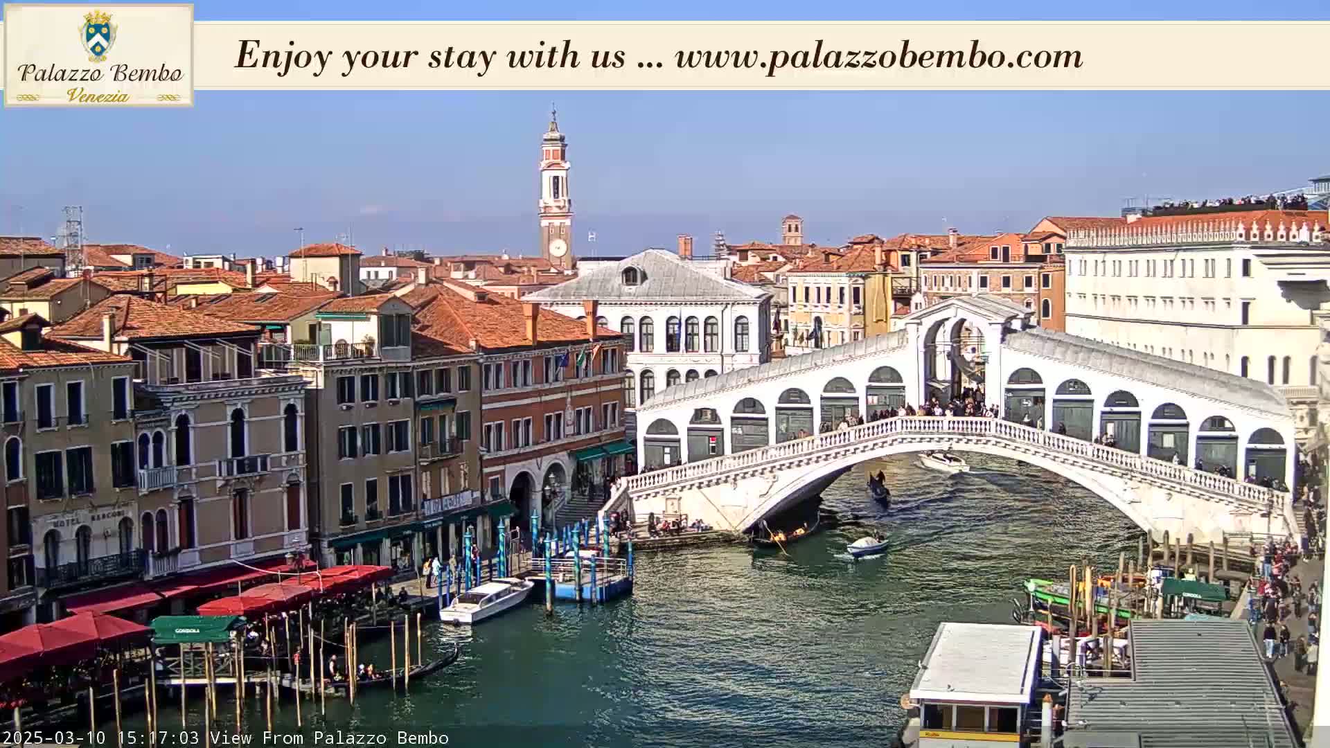 A sunny day in Venice shows a canal with gondolas and boats passing under a bridge, with buildings lining the waterway.