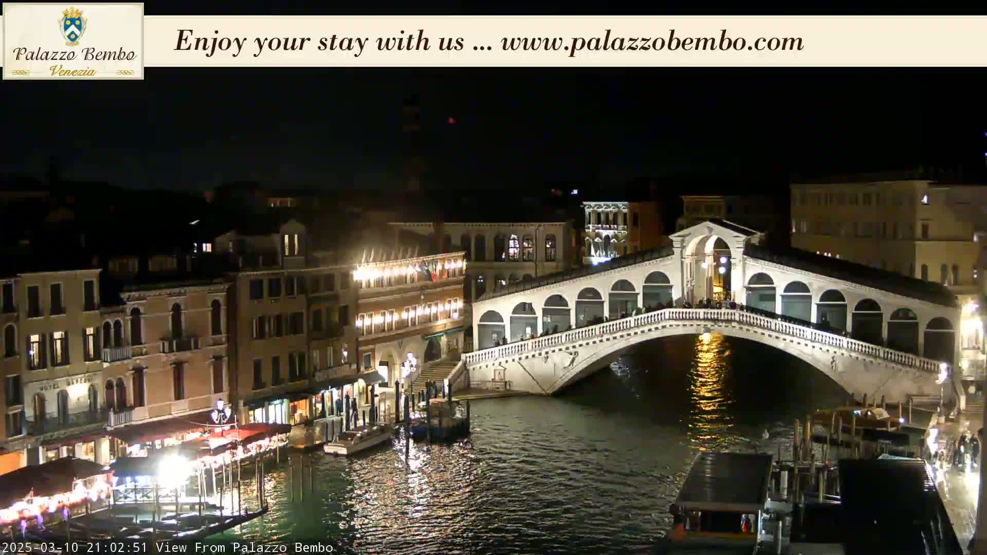 A nighttime view of a bridge spanning a canal in Venice, with buildings illuminated along the waterway.