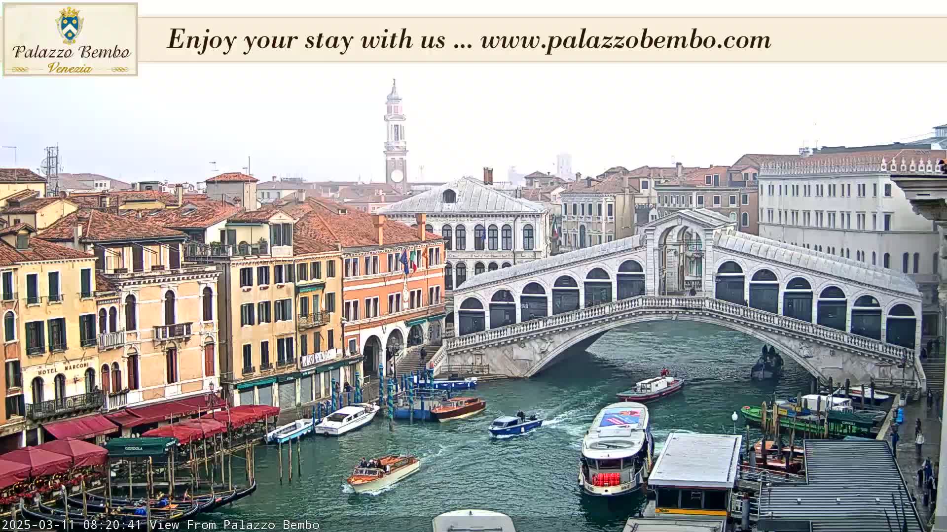 A view of a canal in Venice, Italy, on an overcast day, showing numerous boats and the Rialto Bridge.