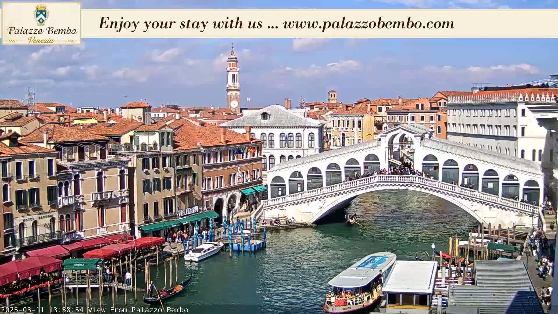 A sunny view of a canal in Venice, Italy, shows a bridge crowded with people, boats, and buildings lining the waterways.